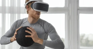 Vyras sportuoja naudodamas virtualios realybės akinius ir kamuolį, sporto salėje su dideliais langais ir natūraliu apšvietimu.